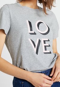 Šedé tričko s krátkým rukávem s nápisem "LOVE" v různých stylech písma; růžové a černé písmeno na měkké, texturované tkanině.