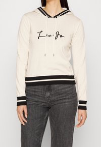 Sweat à capuche couleur crème avec accents noirs, doté d'un cordon de serrage, de poignets et d'un ourlet inférieur côtelés, avec le texte "Liu Jo" en cursive à l'avant.