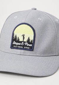 Căciulă de baseball gri deschis cu un patch brodat care afișează un jucător de golf în fața copacilor și a răsăritului de soare. Are un voal curbat și un spate ajustabil.
