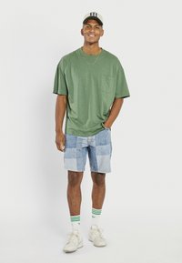 Green short-sleeve t-shirt with pocket, blue patchwork denim shorts, white socks with green stripes, cream sneakers, and a beige cap.