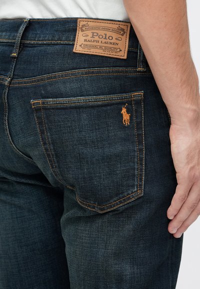 Jeans de mezclilla azul oscuro con costuras naranjas, parche de cuero de la marca Polo Ralph Lauren encima del bolsillo derecho y logo bordado en el bolsillo.
