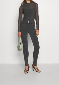 Czarny siatkowy top z długim rękawem w wzór, szare dopasowane jeansy typu skinny z wysokim stanem, zielona torebka oraz czarne sandały na wysokim obcasie z paskami.