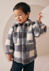 Un jeune enfant souriant portant une veste moelleuse à carreaux gris et blanc, tenant un objet blanc courbé contre un fond beige.