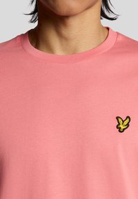 T-shirt in cotone rosa con scollo rotondo e un piccolo logo di un uccellino ricamato in giallo sul lato sinistro del petto. Texture liscia e leggera.