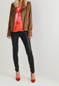 Giacca da motociclista in suede marrone con cerniere argentate, blusa floreale rosa vivace, pantaloni in ecopelle neri e décolleté rossi a spillo.