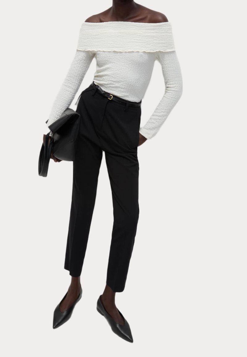Personne portant un top blanc texturé à épaules dénudées et manches longues, un pantalon noir taille haute, des ballerines pointues noires, et tenant une pochette noire.