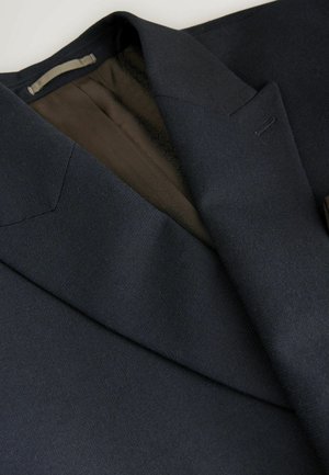 Blazer marine en tissu texturé. Il présente un col cranté, des coutures discrètes et une doublure à motifs bruns visible à l'intérieur.