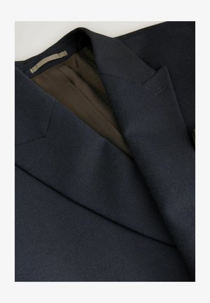 Blazer marine en tissu texturé. Il présente un col cranté, des coutures discrètes et une doublure à motifs bruns visible à l'intérieur.