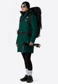 Green waterproof jacket with a cinched waist, black backpack, and trekking poles. Accessories include sunglasses and a beanie. Black leggings and shoes.