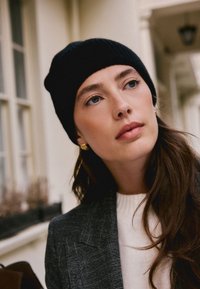 Bonnet noir en tricot, blazer gris à motif, et pull blanc à côtes. Boucles d'oreilles circulaires en or et cheveux bruns ondulés visibles.