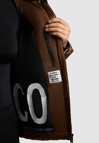 Giacca imbottita marrone con rivestimento nero, chiusura con zip e un'etichetta che dice "Un abbraccio caldo fatto di tessuto." Presenta un grande testo bianco all'interno.