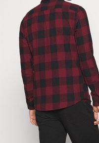 Chemise à manches longues à carreaux rouges et noirs, avec un col boutonné et un ourlet arrondi. Fabriquée en tissu doux avec une texture lisse.