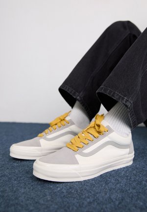 Gray and white sneakers with yellow laces, suede and canvas material, low-top design, and a textured sole, worn with dark jeans and white socks.