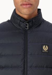 Navy blue puffer jacket with a high collar, zip closure, and small gold logo patch. Features a quilted texture and sleek design.