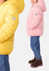 Two puffer jackets shown: one yellow with a hood and logo on the sleeve, the other pink, both featuring quilted patterns. Brown boots worn.