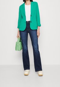 Blazer vert, chemise blanche, jeans évasés bleu foncé, sabots crème et un sac à main vert clair. Tissu lisse, design ajusté et couleurs contrastantes.