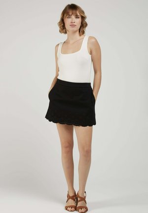 Femme debout, les mains dans les poches, portant un haut blanc sans manches, une jupe noire à ourlet festonné et des sandales à talons marron sur un fond uni.