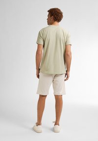 Homme aux cheveux bouclés portant une chemise à manches courtes vert clair, un short beige jusqu'aux genoux et des baskets blanches, se tenant de dos sur un fond blanc.