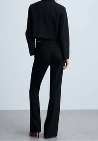 Blazer noir court associé à un pantalon noir ajusté. La tenue présente une texture lisse et un design structuré. Des talons rouges soulignent le look.