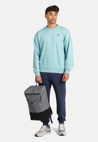 Sudadera azul claro con puños acanalados, mochila gris y negra con diseño geométrico, y zapatillas grises con acentos, de pie contra un fondo blanco.