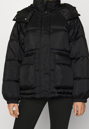 Schwarze Steppjacke mit hohem Kragen, zwei Klappentaschen, elastischen Bündchen und abnehmbarer Schulterklappe, getragen über schwarzen Hosen.