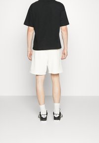 Mann mit schwarzem T-Shirt, weißen Shorts, weißen Socken und schwarzen Adidas-Sneakern, der vor einem schlichten weißen Hintergrund steht.