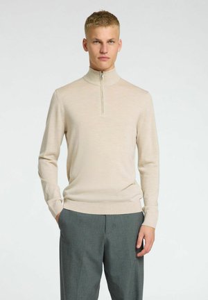 Beige zip-neck genser i en jevn, lett strikk, med lange ermer og en tettsittende design. Kombinert med grå, skreddersydde bukser.