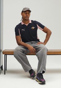 Juodas polo marškinėlis su rožiniais akcentais, šviesiai pilkos džinsinės kelnės ir margaspalviai sportbačiai. Modelis sėdi ant medinės suolelio prieš baltą foną.