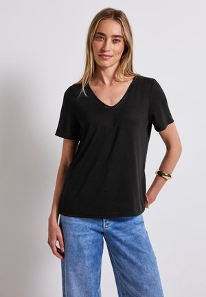 Zwarte V-hals T-shirt gemaakt van zacht stof. Korte mouwen, een relaxte pasvorm, met kleine uitsnijdingen langs de decolleté. Gecombineerd met lichtblauwe spijkerbroek.