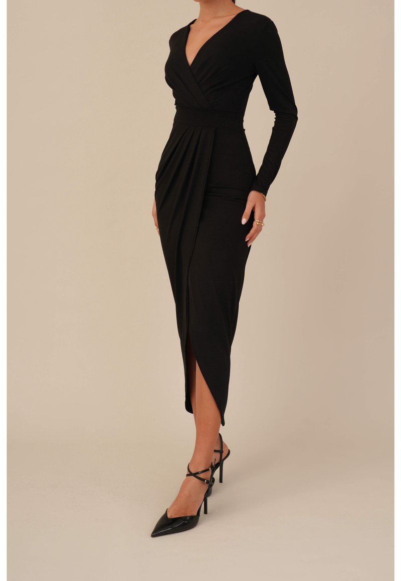 Femme portant une robe midi noire moulante à manches longues avec un devant croisé et une fente avant, associée à des escarpins noirs à brides.