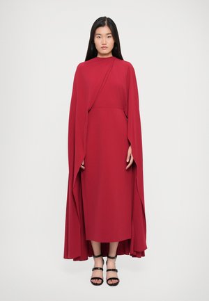 Vrouw staat in een rode full-length jurk met een aanraakbare vloeiende cape en zwarte bandjes met hoge hakken op een witte achtergrond.