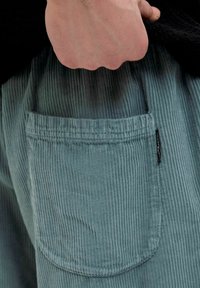Pantaloni din corduroy de culoare albastru deschis, cu o textură specială, având un buzunar aplicat și o etichetă neagră pe lateral, subliniind designul lor casual.