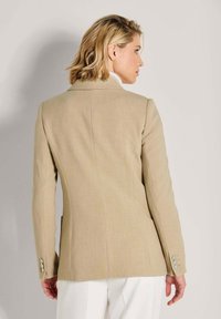 Vrouw met kort blond haar, gekleed in een beige op maat gemaakte blazer en witte broek, gezien vanuit de rug tegen een effen achtergrond.