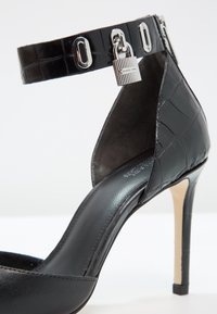 Chaussure à talon haut noire avec texture crocodile, bride de cheville avec accessoires argentés et cadenas, design à bout ouvert, intérieur lisse.