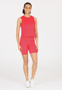 Rode atletische tanktop en bijpassende fitted shorts gemaakt van gladde, rekbare stof. Model draagt witte sneakers en staat op een effen achtergrond.