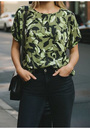 Femme portant un haut vert à manches courtes à motifs, rentré dans un jean noir taille haute, debout avec les mains dans les poches dans une rue de la ville.