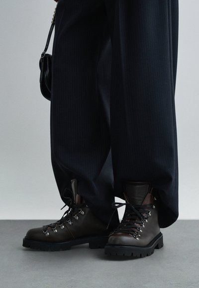 Stivali da trekking in pelle marrone con suola in gomma, occhielli in metallo e lacci neri. Abbinati a pantaloni larghi a righe sottili scuri.