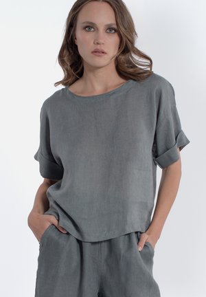 Donna che indossa una maglietta grigia a maniche corte e pantaloni abbinati, mani nelle tasche, guarda avanti con espressione neutra su sfondo semplice.
