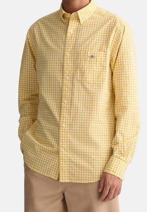 Homme portant une chemise à carreaux jaune et blanche à manches longues avec poche poitrine et pantalon beige, debout les mains détendues.