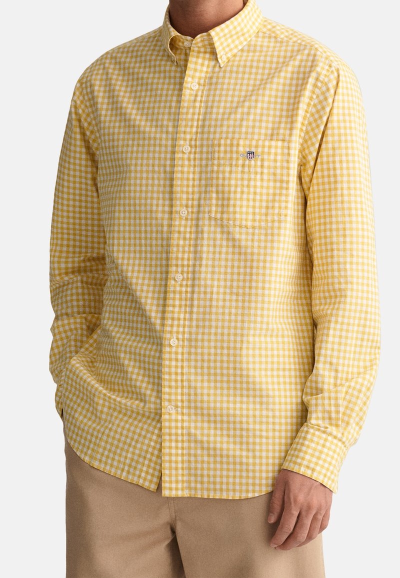 Homme portant une chemise à carreaux jaune et blanche à manches longues avec poche poitrine et pantalon beige, debout les mains détendues.