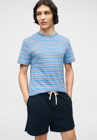 Νεαρό άτομο με κοντά σκούρα μαλλιά φοράει μπλε ριγέ μπλούζα και σκούρα σορτς με κορδόνι, στέκεται με τα χέρια στις τσέπες.