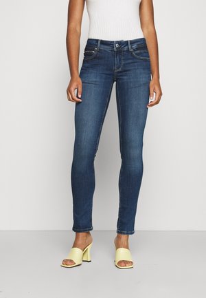 Femme portant un jean skinny bleu foncé, un haut sans manches blanc et des chaussures ouvertes à talons jaunes, debout sur un fond uni.