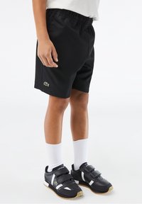Enfant portant un short de sport noir avec un petit logo vert, des chaussettes blanches et des baskets noires à scratchs, debout sur un fond blanc.