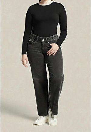 Personne portant un haut noir à manches longues, un jean taille haute gris foncé et des baskets blanches, debout devant un fond uni.