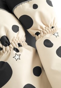 Des mitaines beiges avec de grands pois noirs, des poignets élastiques et un petit détail graphique en forme d'étoile. Le matériau semble lisse avec une finition légèrement brillante.