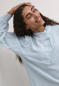 Blouse bleu clair avec un col volanté et un plastron à boutons. Manches longues avec des poignets froncés, fabriquée à partir d'un tissu lisse et texturé.