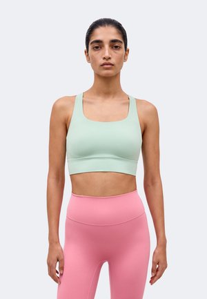 Γυναίκα με μαλλιά δεμένα πίσω, φοράει αθλητικό μπουστάκι σε χρώμα μέντας και ροζ κολάν με ψηλή μέση, στέκεται σε ένα απλό φωτεινό φόντο.