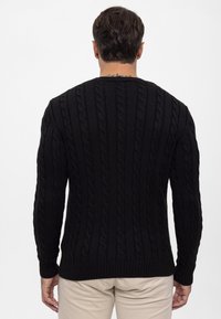 Pull en tricot noir à motif texturé en relief, avec des poignets et un ourlet côtelés. Le design présente des motifs de câbles verticaux sur l'ensemble.