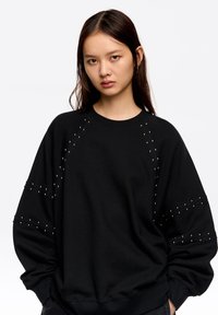 Sudadera negra con cuello redondo, mangas largas y detalles de tachuelas en los hombros y las mangas, confeccionada con un tejido suave.