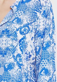 Blusa blu e bianca con motivo floreale e forme geometriche, realizzata in tessuto leggero e con colletto.